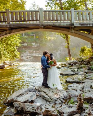 Newlyweds share an intimate moment on rocky riverbank under an arched bridge.