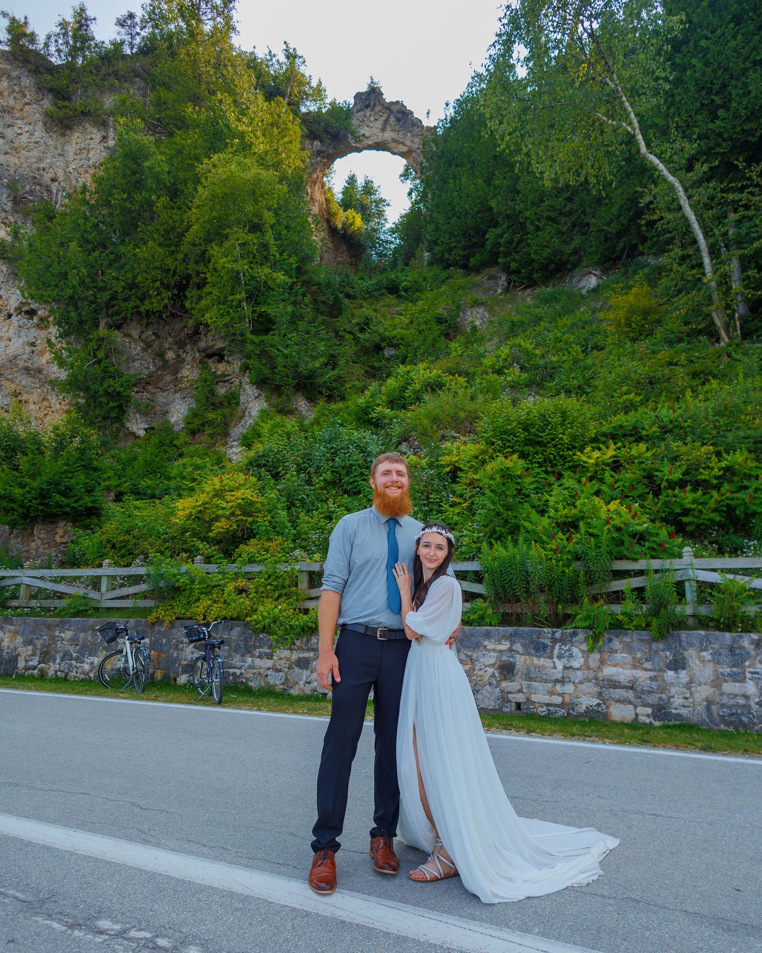 A scenic wedding photo taken in front of a natural stone arch formation surrounded by lush greenery.