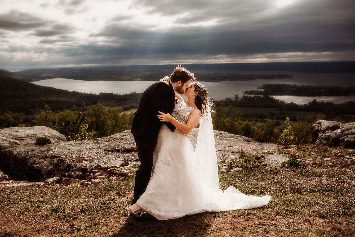 Dramatic wedding photos on a clifftop overlooking a scenic mountain landscape at sunset.
