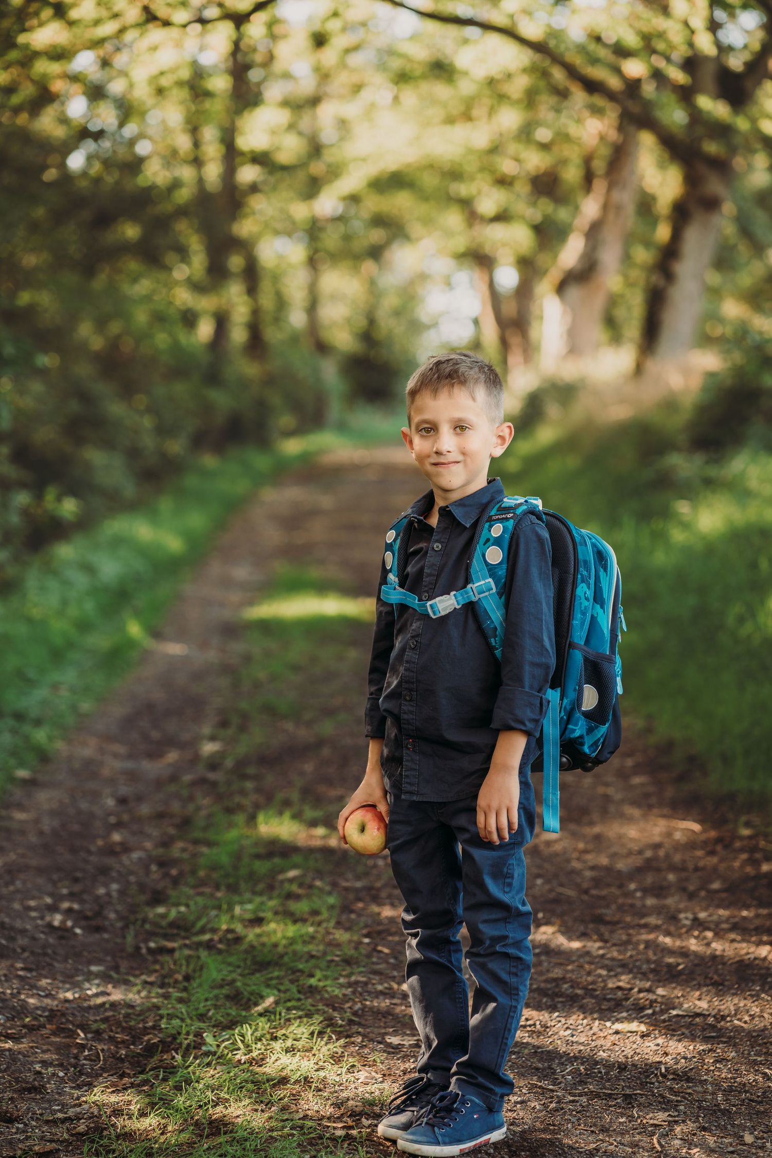 Student wearing blue backpack standing on forest path ready for school.