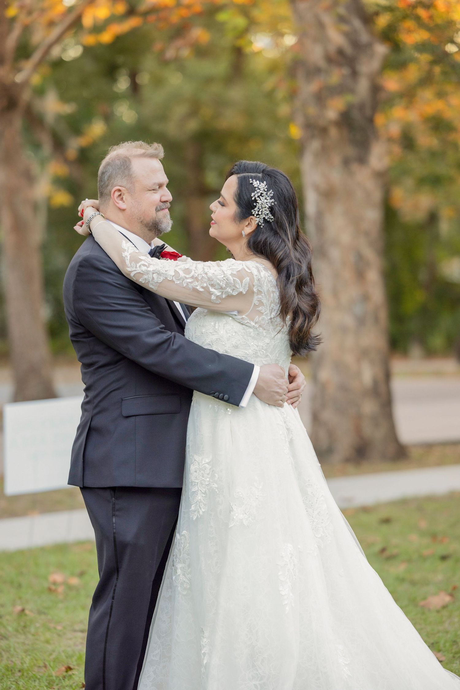 A couple embraces outdoors in wedding attire surrounded by autumn trees with golden leaves in the background.