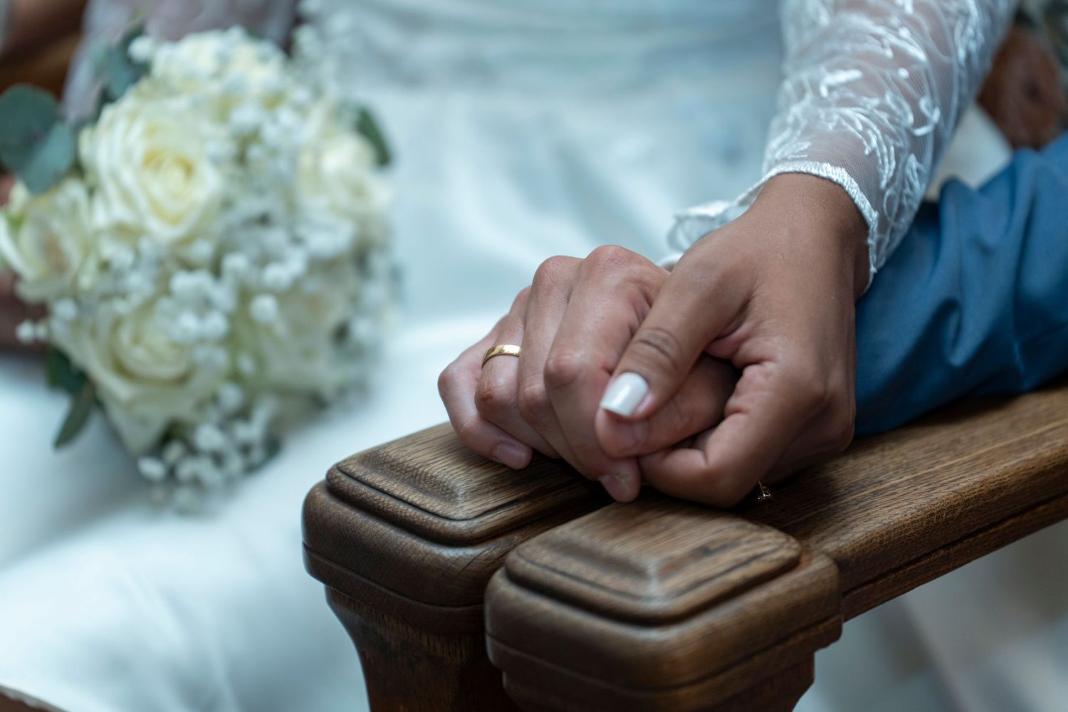 Hands resting on wooden armrest with white lace sleeve and wedding bouquet in soft focus background.