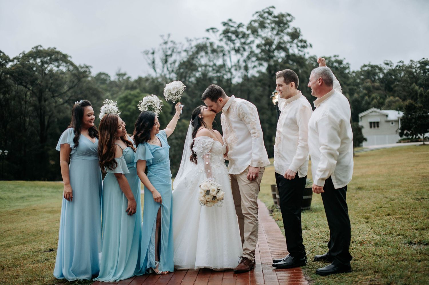 Wedding party in light blue and white attire gathered on a wooden platform outdoors.