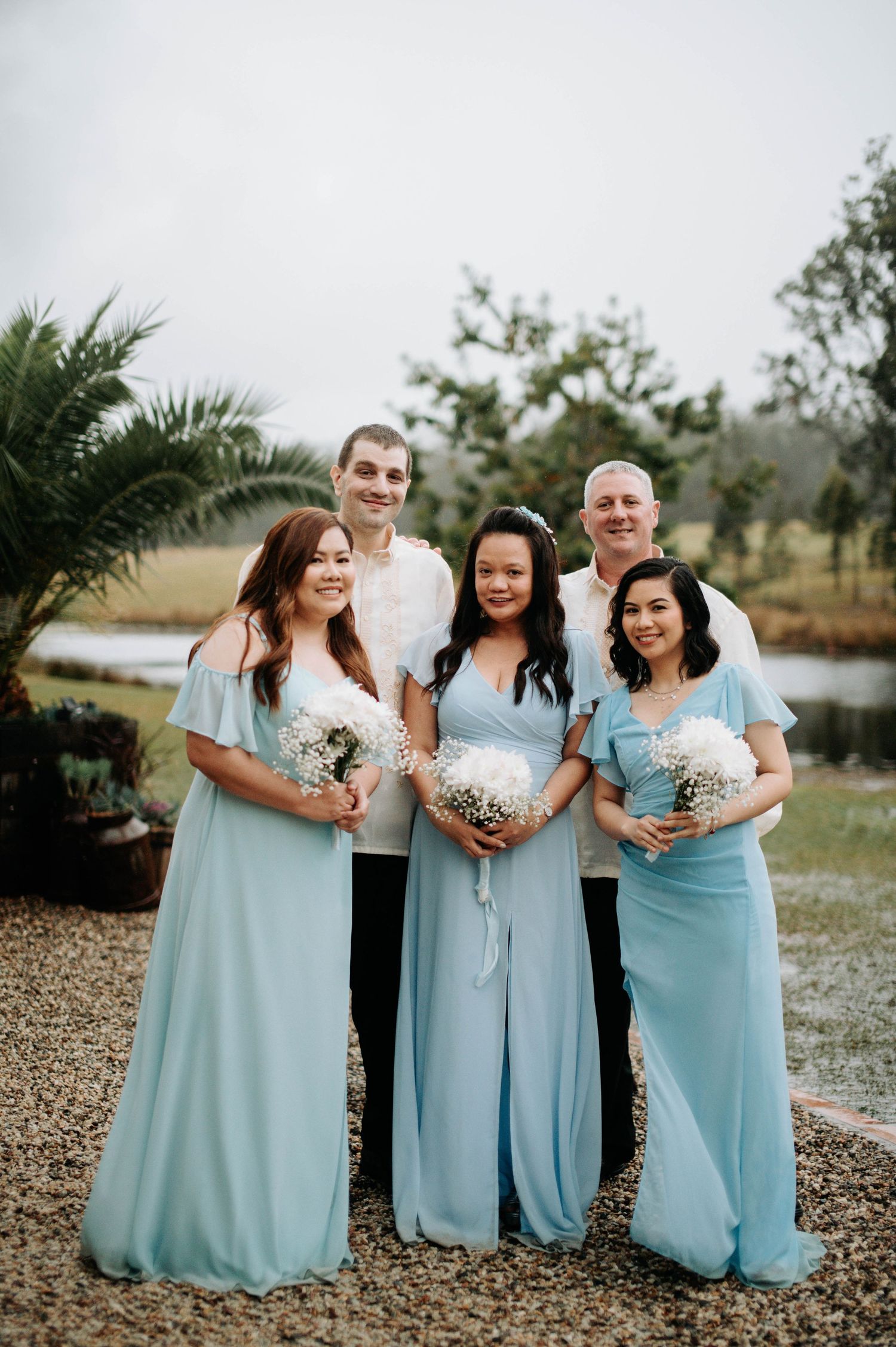 Wedding party group photo in coordinated light blue and white attire by a pond.