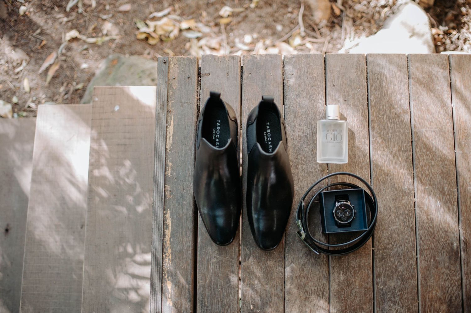 Black formal shoes and a ring box arranged on wooden decking in natural light.