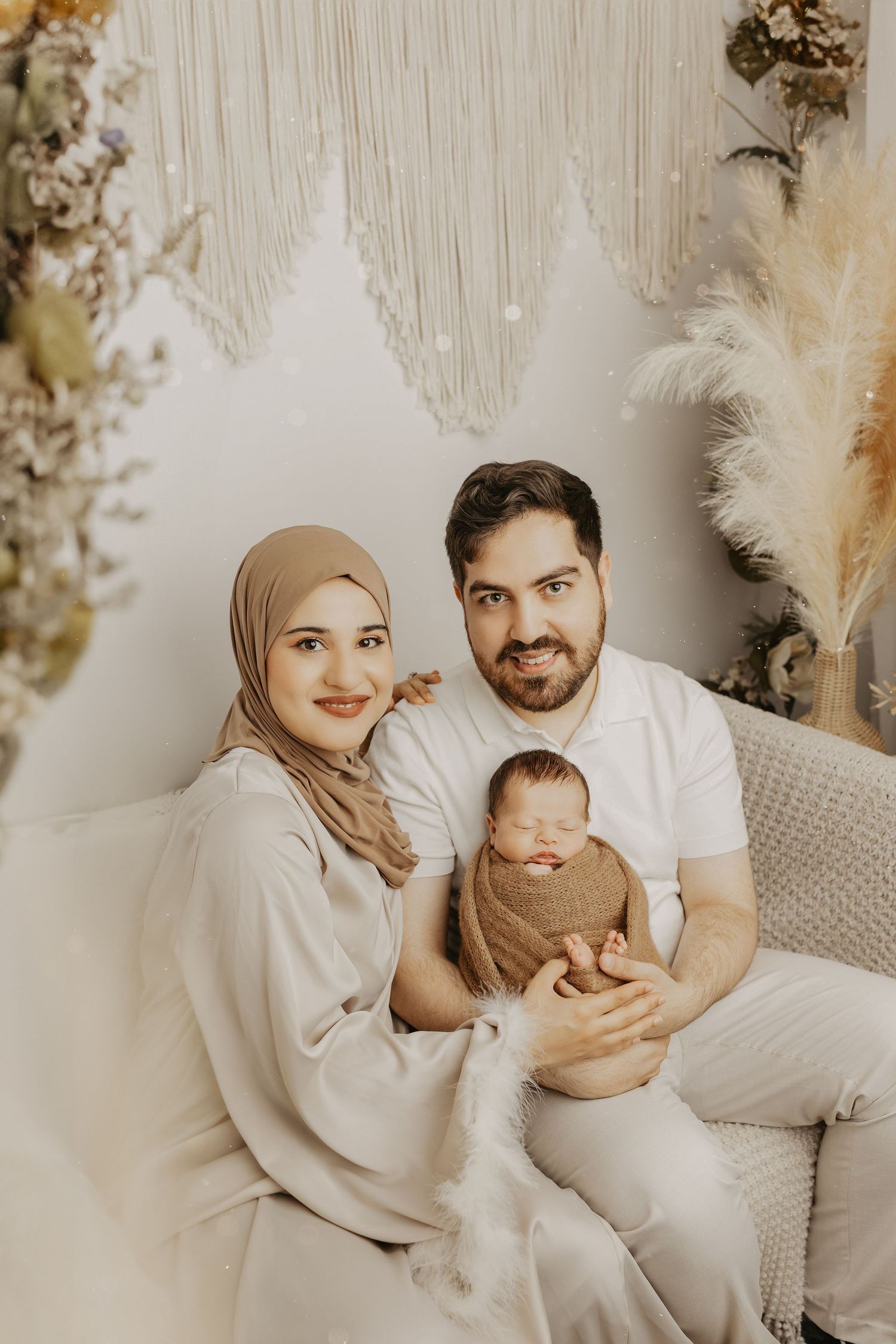 Family portrait featuring new parents with baby in neutral tones against floral backdrop.
