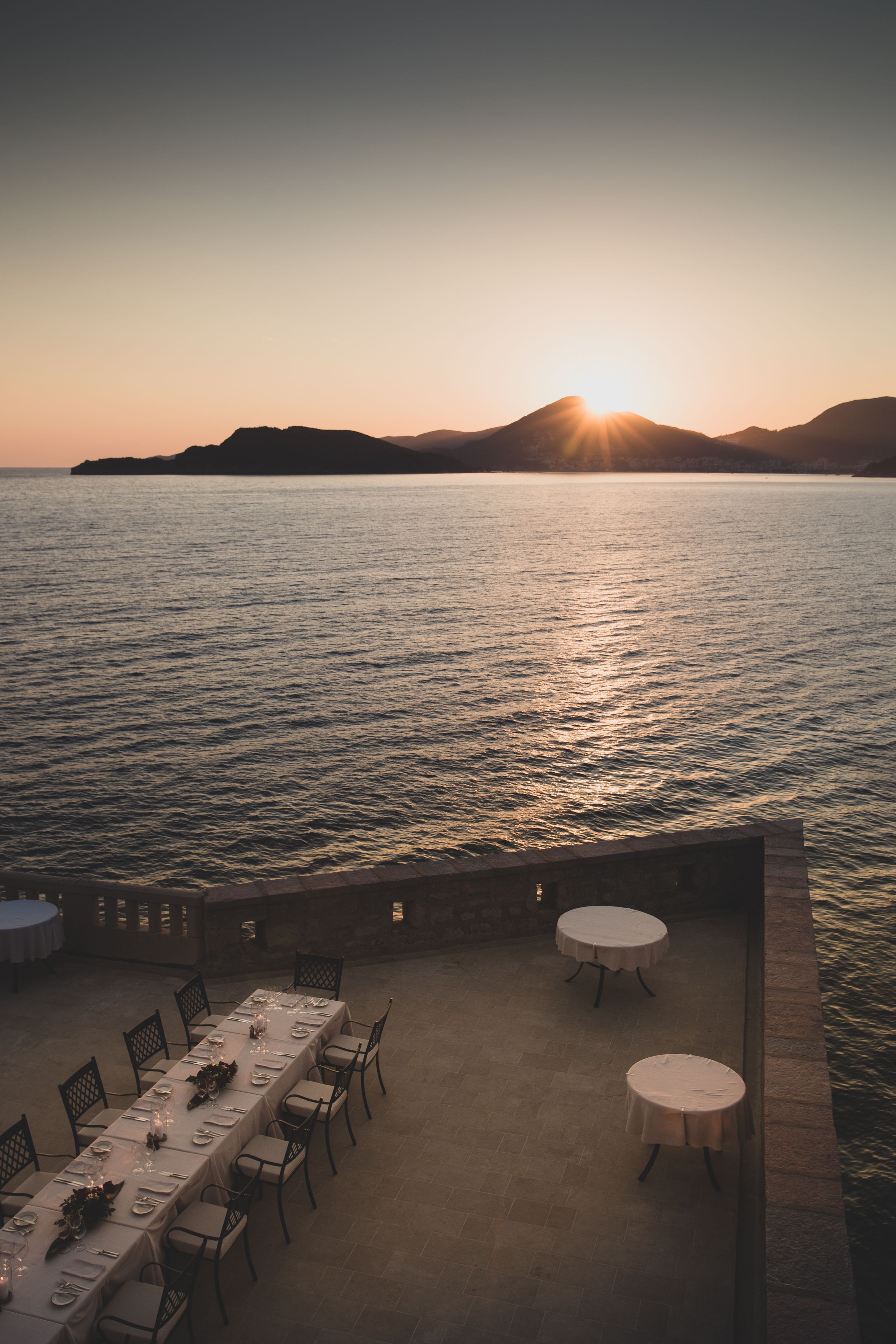Sunset dining terrace overlooking ocean with empty tables and mountain silhouettes.