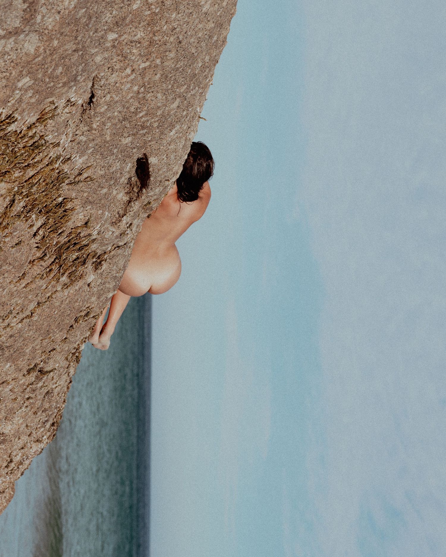 Artistic nude photograph of a figure climbing a rock face against a blue sky.