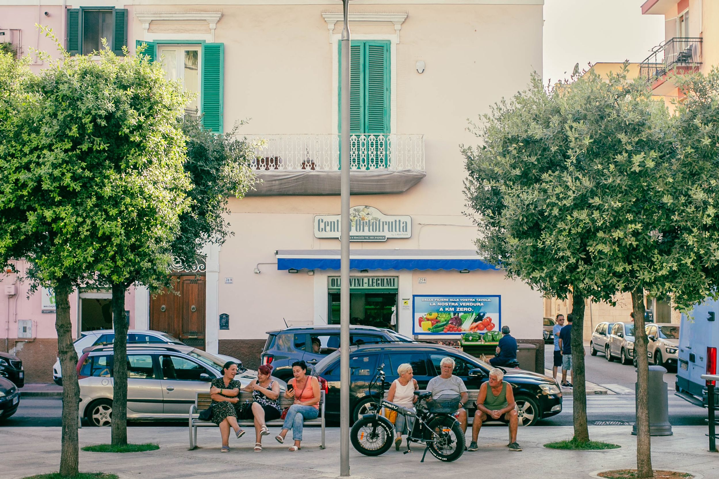 People gathering on a Mediterranean street corner with green trees and parked vehicles.