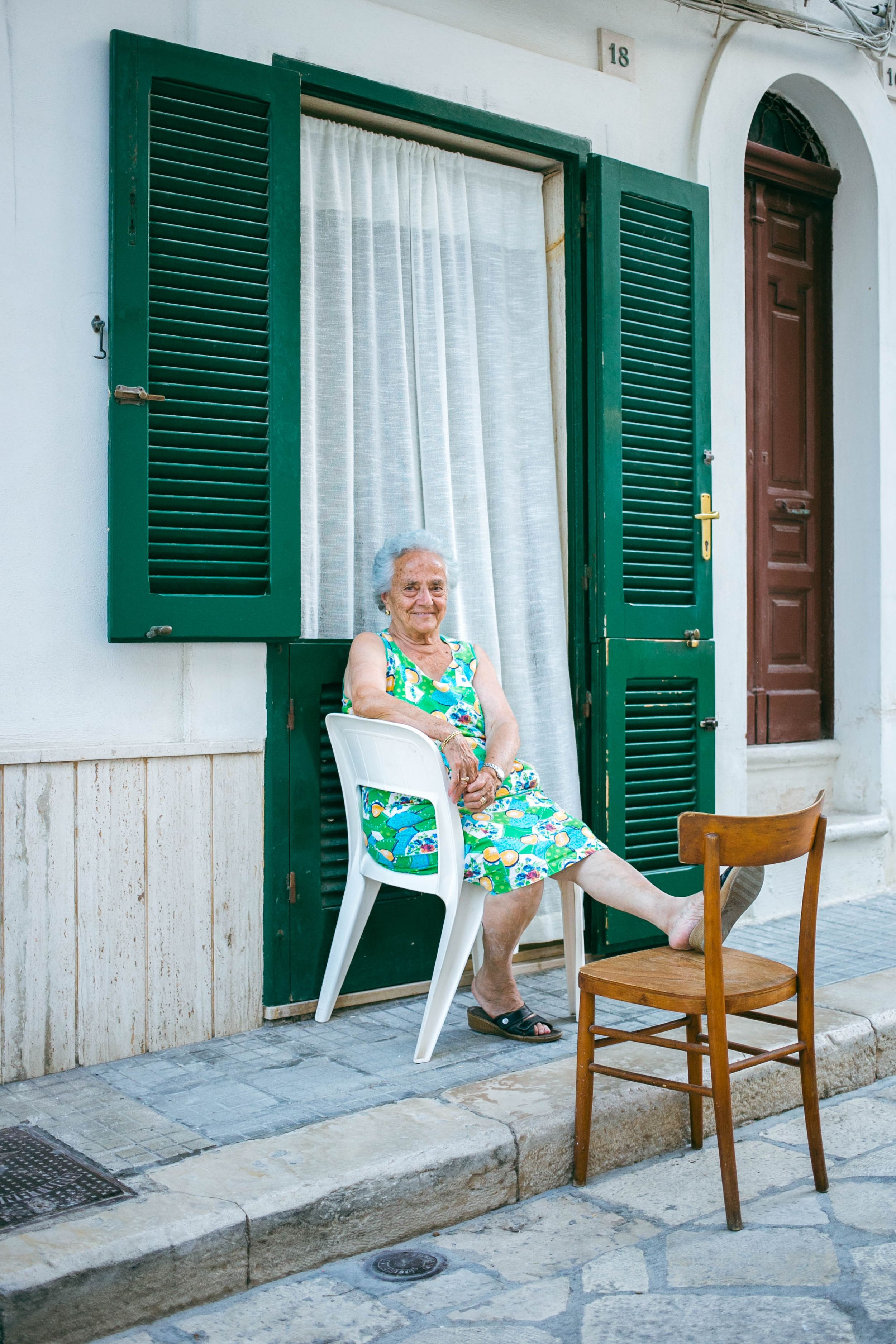 Person sitting in a white chair outside a building with green shutters and a wooden door.