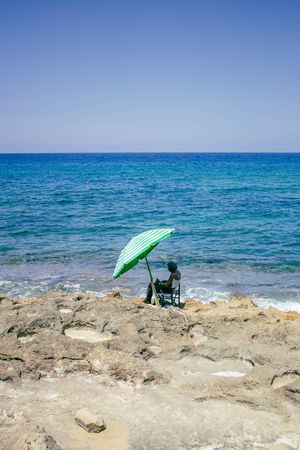 Person sitting under green umbrella on rocky coastline overlooking bright blue Mediterranean sea.