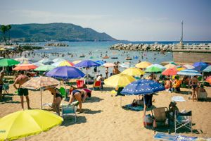 Colorful beach umbrellas dot a crowded Mediterranean beach with mountains in the background.