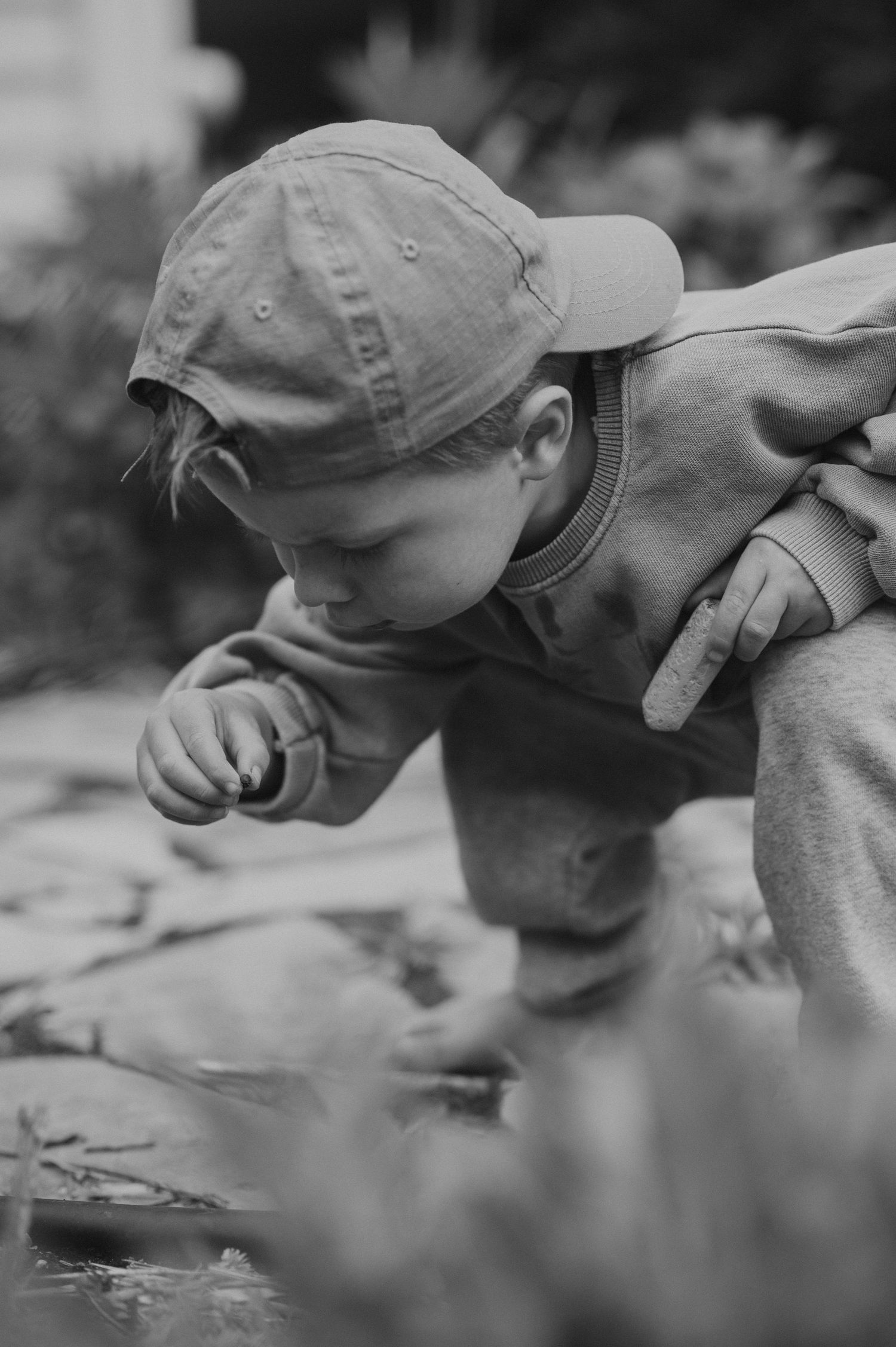 Black and white photo of child in baseball cap leaning over to examine something up close.