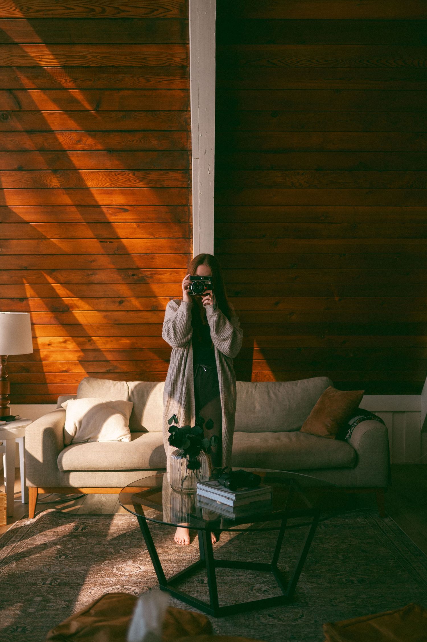 Sunlight streams through blinds onto wood paneling while a photographer takes a picture in a cozy living room.
