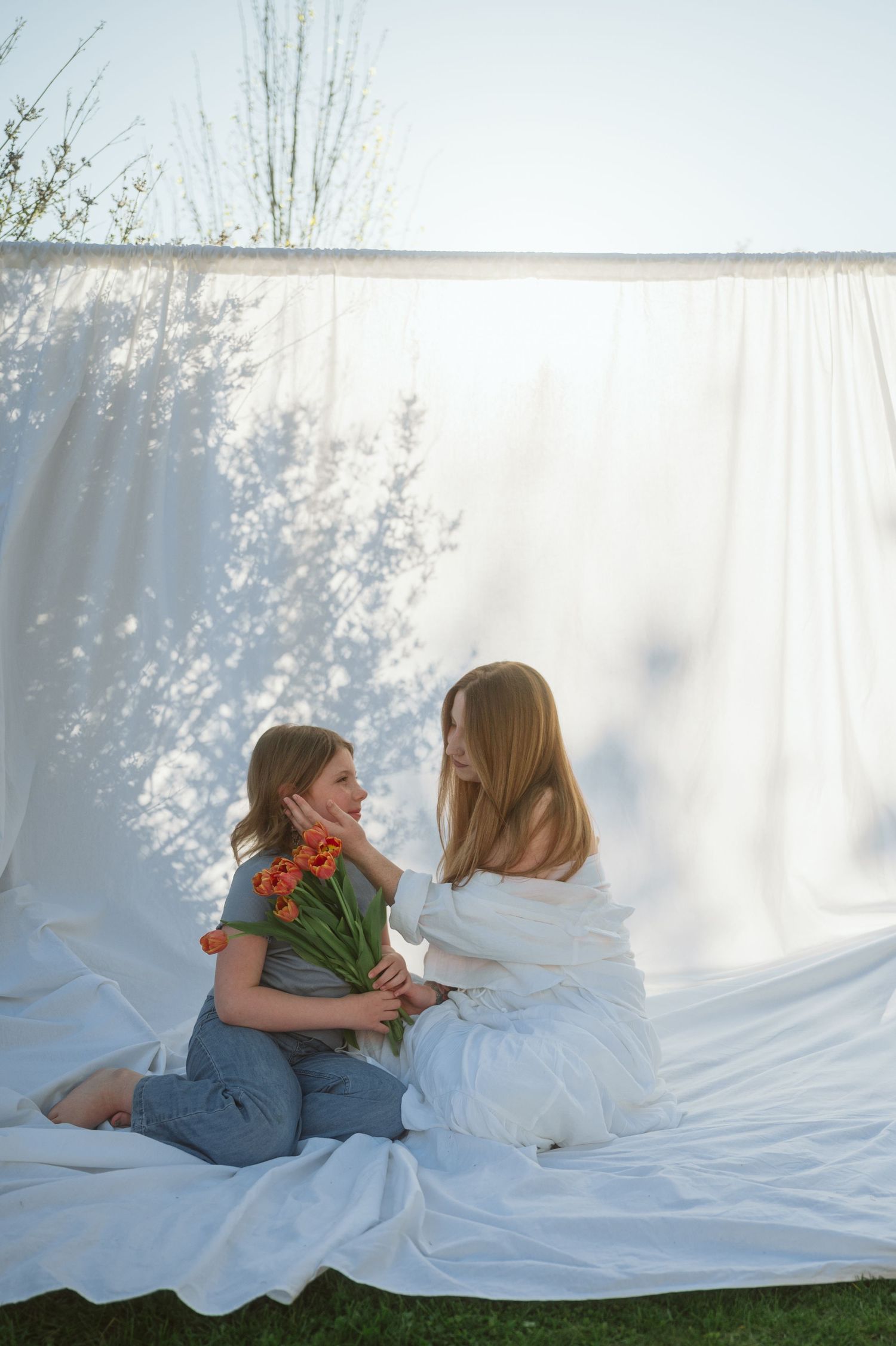 Two people sit on white fabric outdoors holding a bouquet of tulips while delicate tree shadows play on a backdrop.