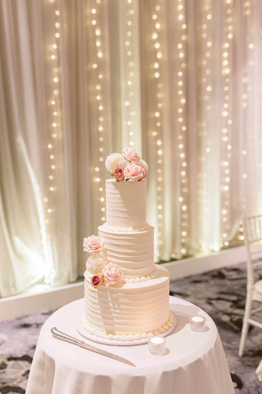 Three-tiered white wedding cake with pink floral decorations against a backdrop of twinkling fairy lights.