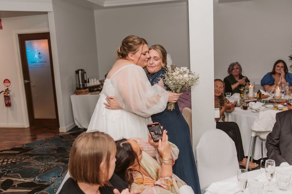 Emotional embrace during wedding reception with guests seated at decorated tables in background.