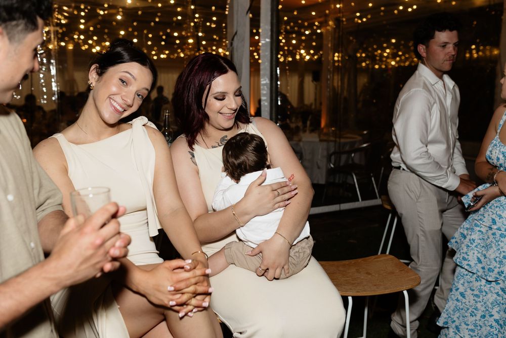 Wedding guests in white dresses share joyful moments at an evening reception with twinkling lights in the background.