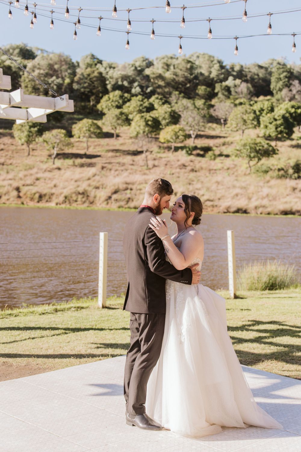 Couple sharing first dance on outdoor dance floor with string lights and lake view.