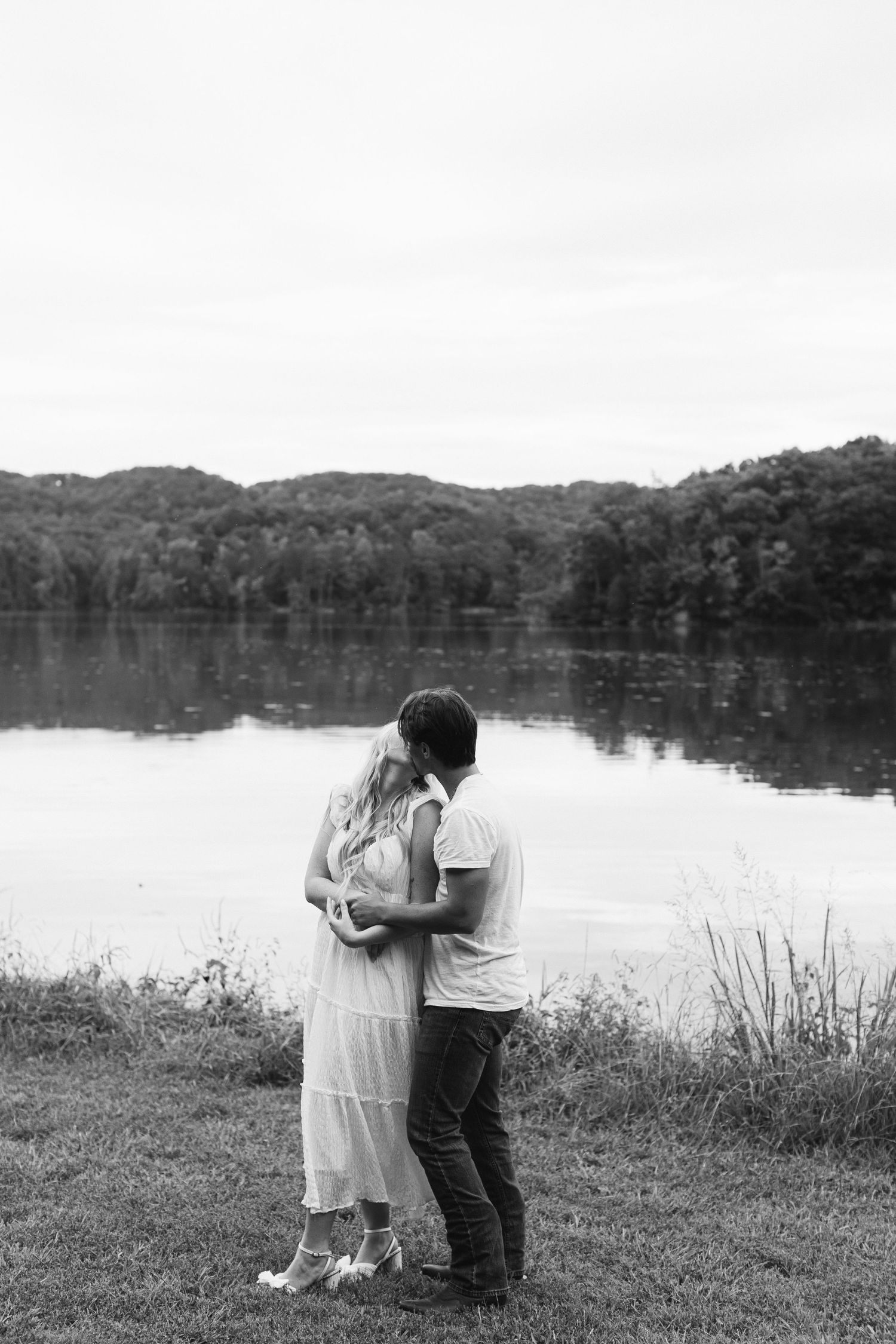 A couple shares an intimate moment by a serene lake in black and white.