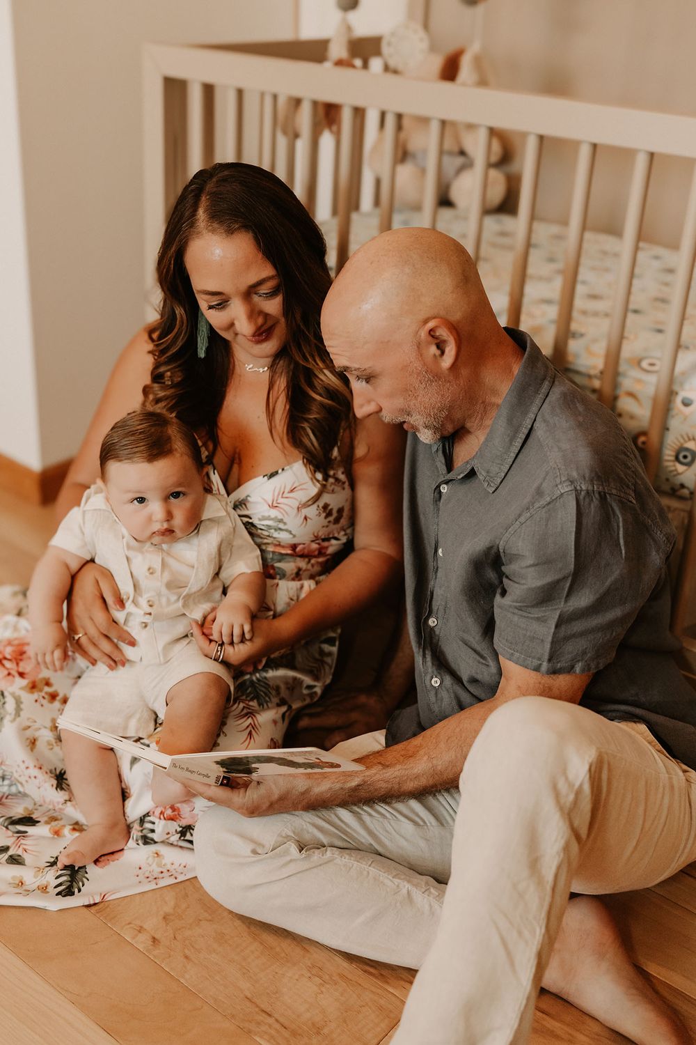 Bald father and brunette mother reading book to baby boy sitting on nursery floor by crib.