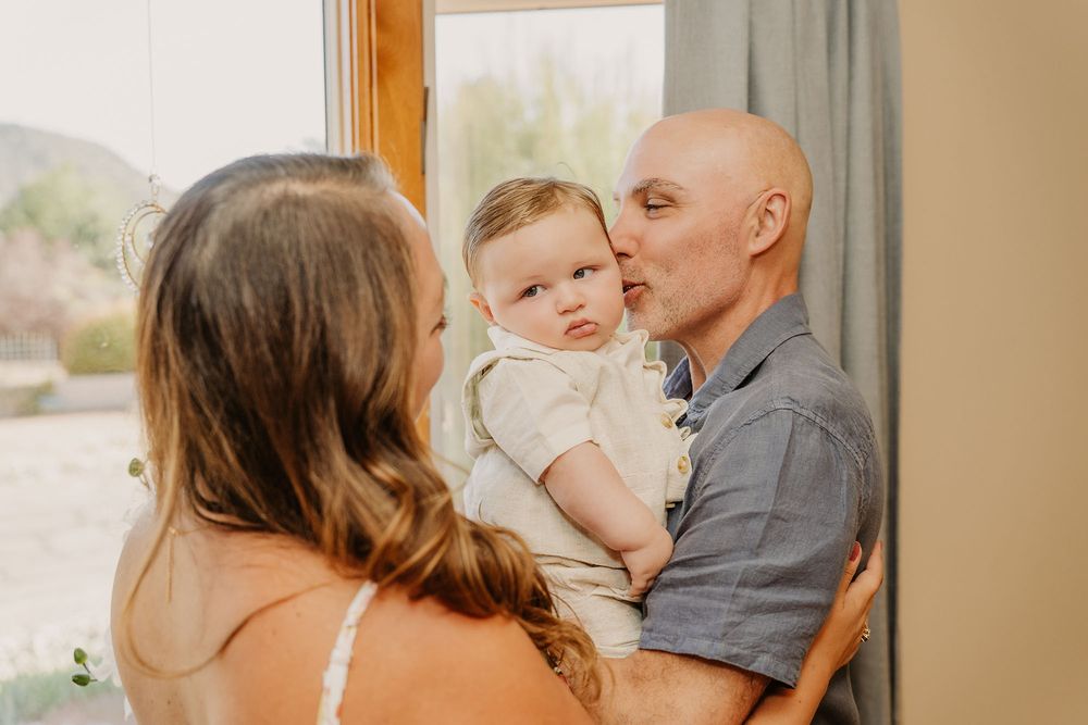 A bald man kisses a baby held in his arms while a woman with long brown hair looks on indoors.