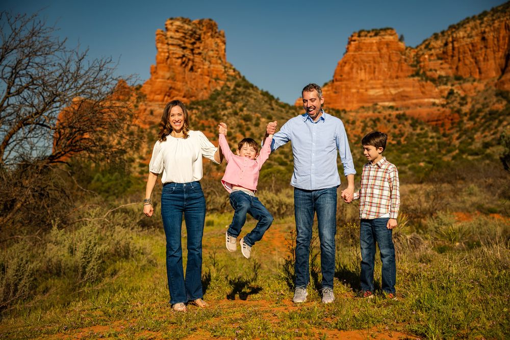 Family jumps joyfully together in front of majestic Sedona red rock formations during outdoor photo session.