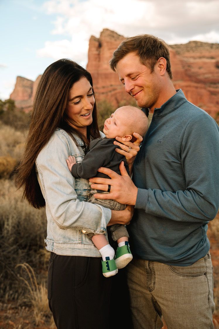 Parents smile lovingly at their infant baby against backdrop of Sedona red rock cliffs during golden hour.
