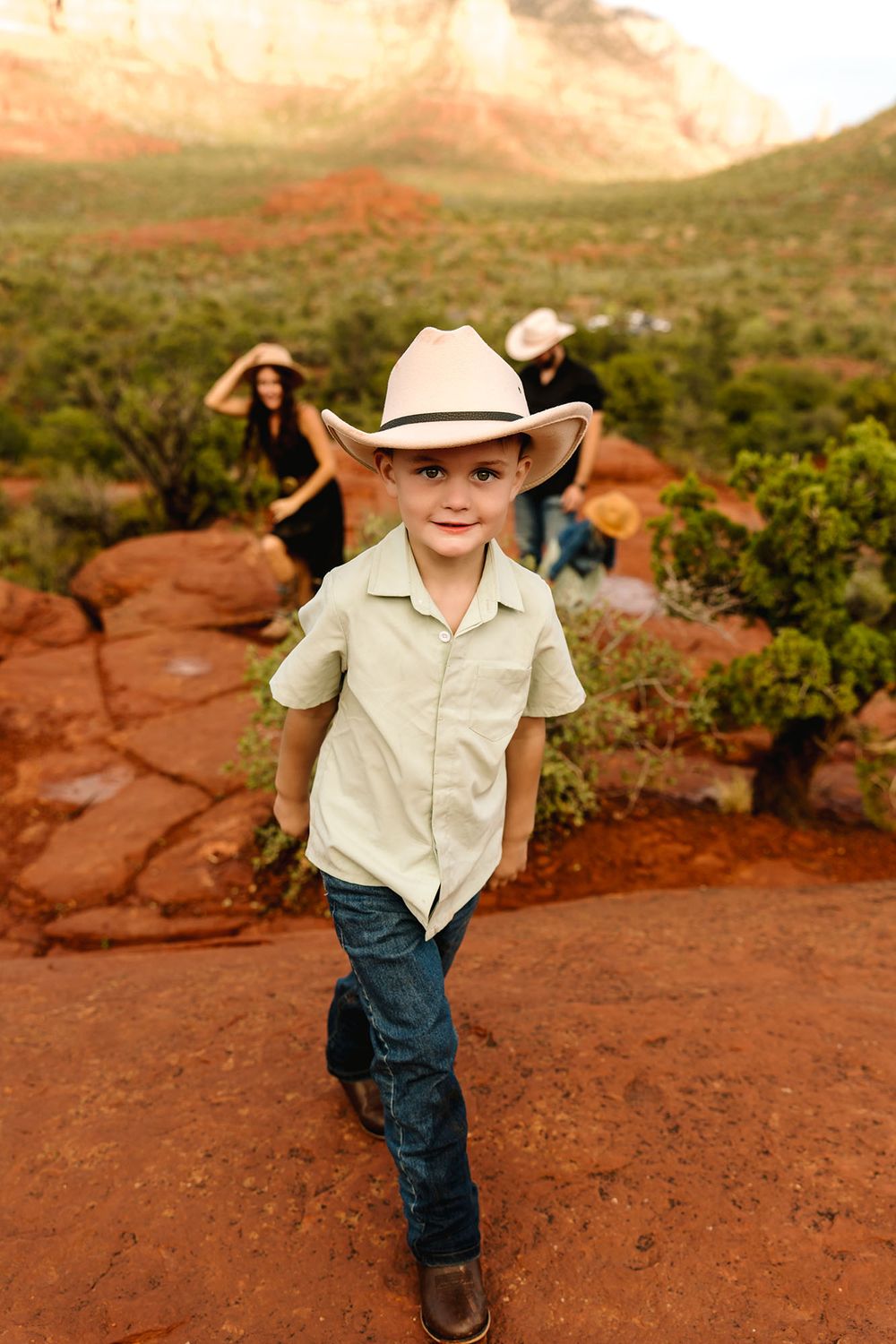 Young boy in cowboy hat and jeans poses on red rocks in Sedona, Arizona, with family members in background.