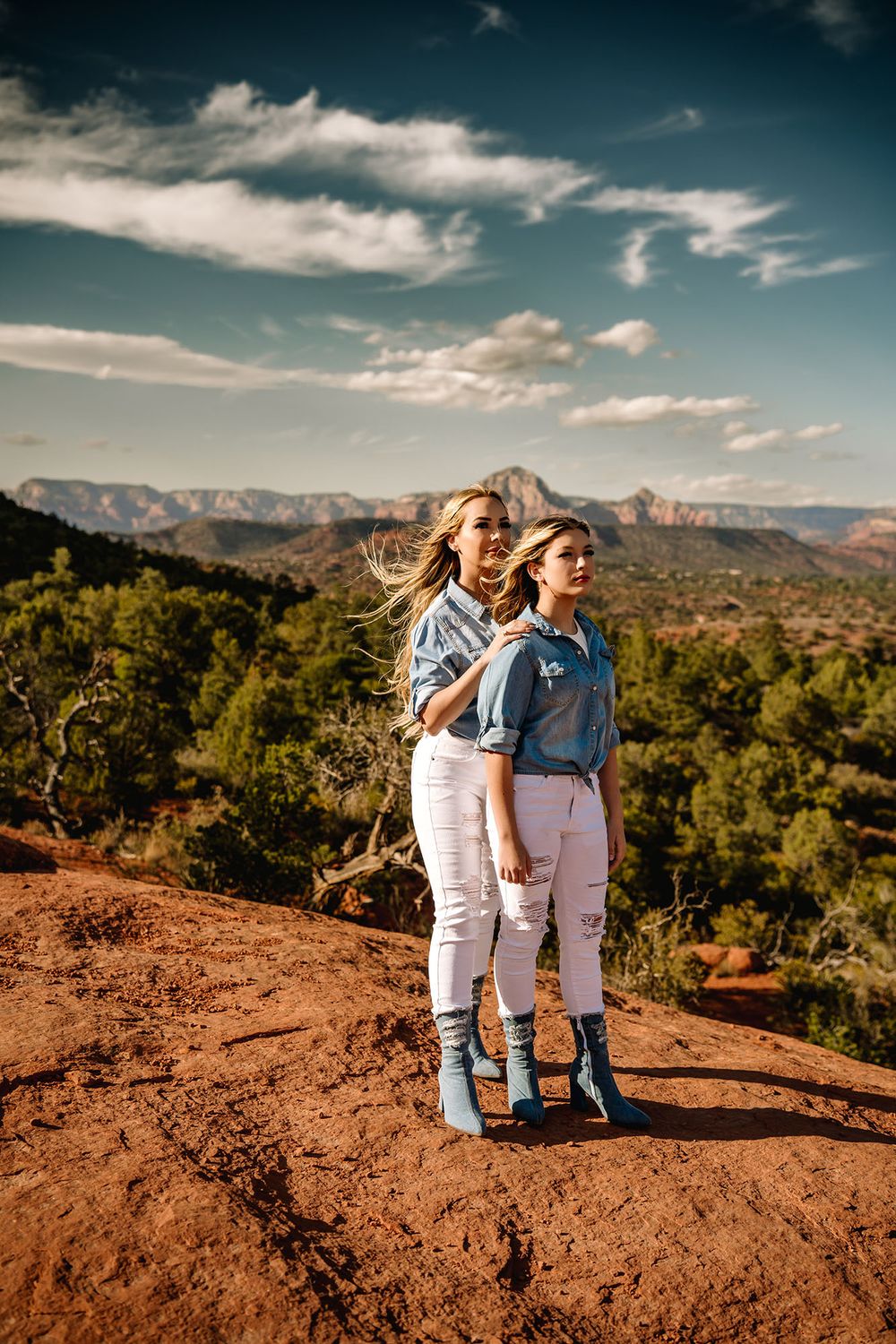 Two women in white jeans and denim shirts stand on red rock with Sedona mountain views.