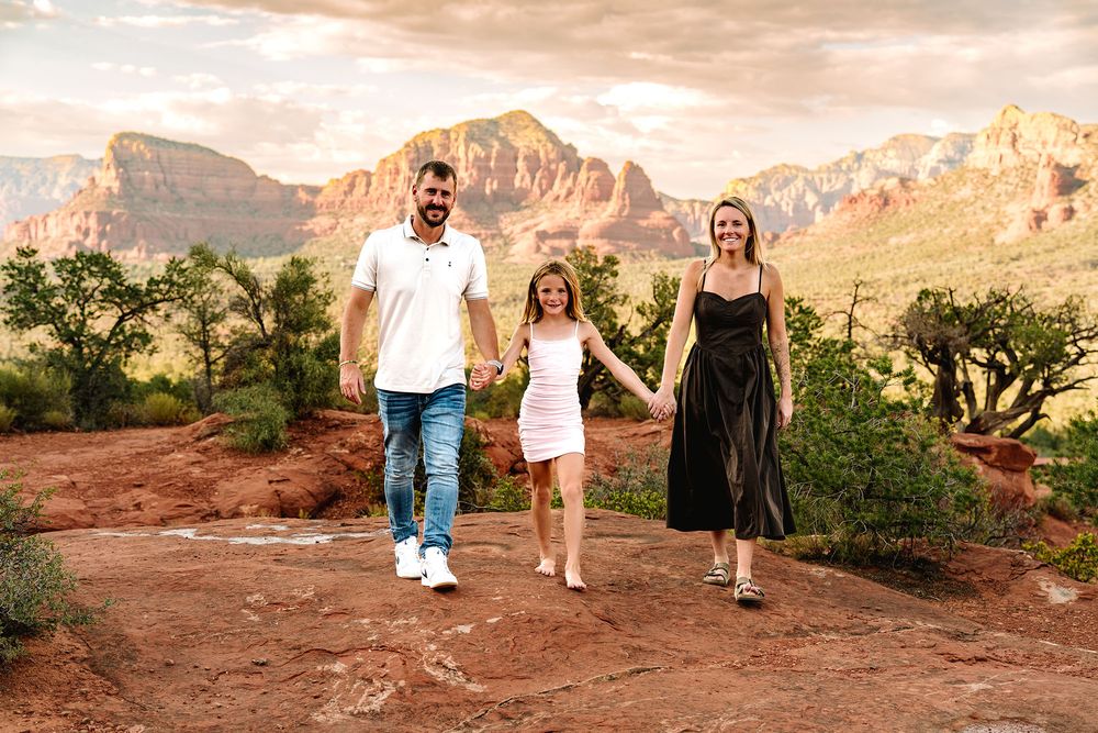 Family of three walks hand-in-hand on red rock trail with stunning Sedona Arizona mountain backdrop at sunset.