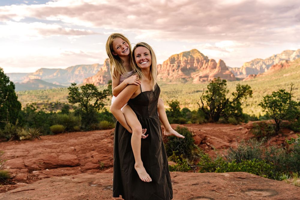 Two women smile at sunset in Sedona, Arizona, with red rock formations and lush greenery in the background.