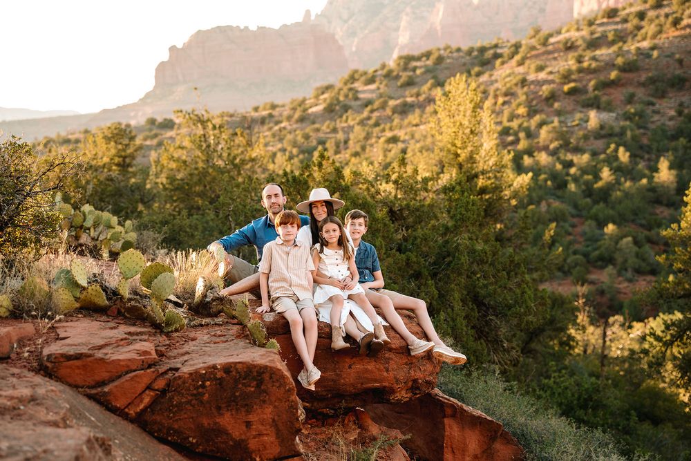 Family of five sitting on red rocks in Sedona Arizona with lush green desert landscape and red cliffs in background.