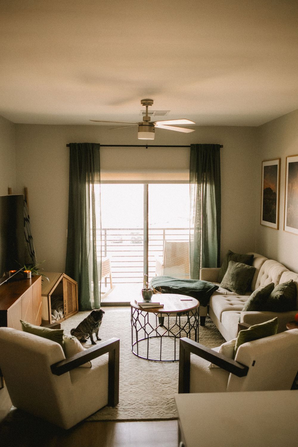 Cozy modern living room with green curtains, white sofa, round coffee table, ceiling fan, and a pug dog sitting on an armchair.