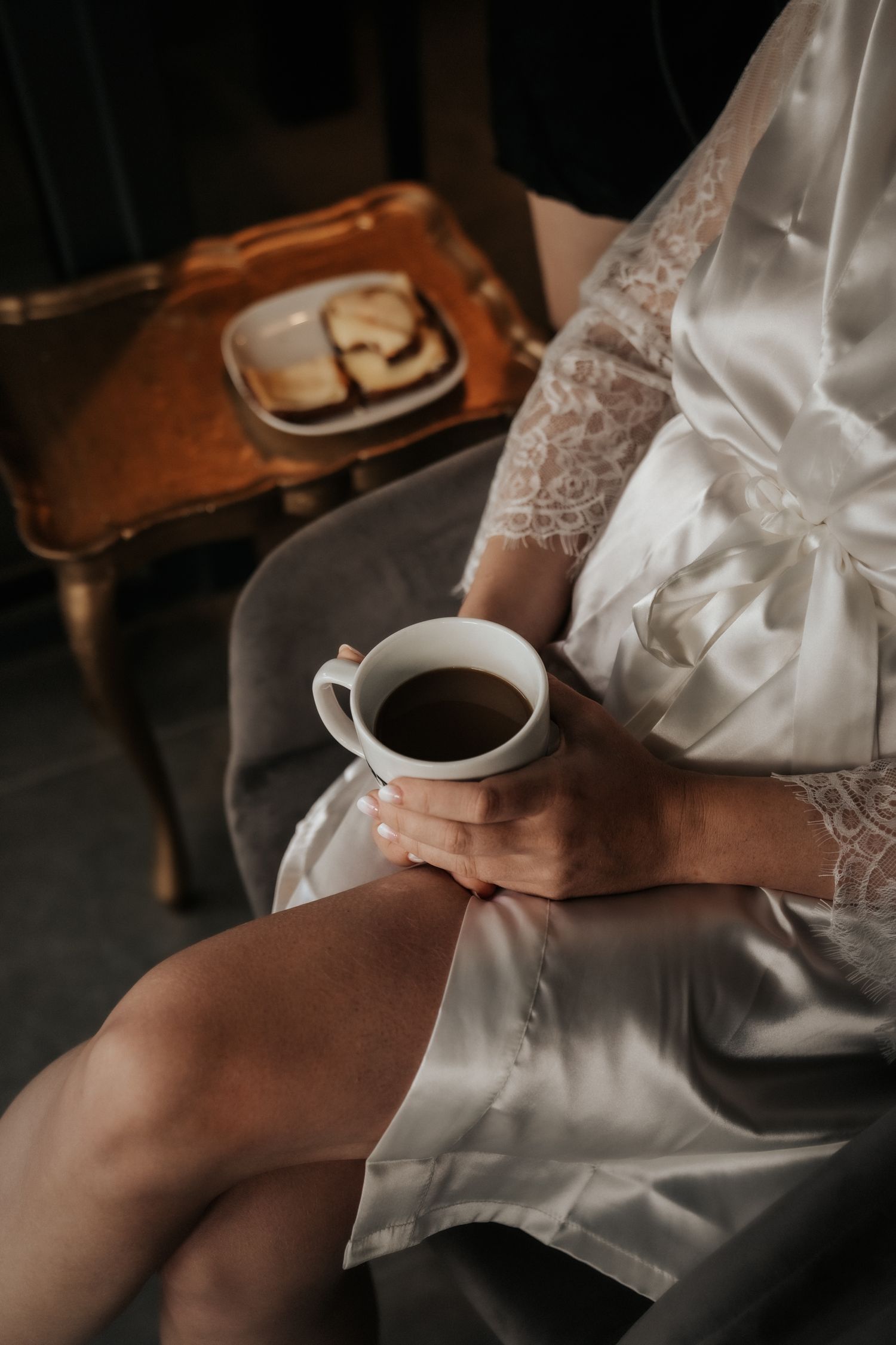 Person in white lace robe holding coffee cup with toast on wooden table in background.