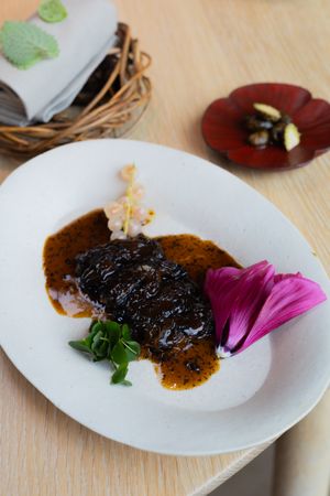 Braised meat in dark sauce garnished with purple flower petals on white plate.
