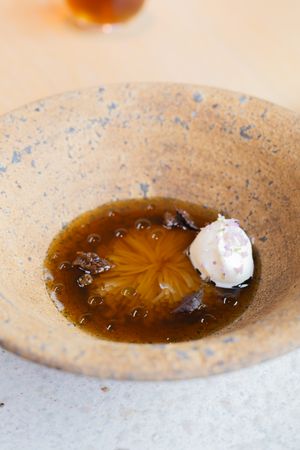 Artistic plating of brown consomme with floral design and white foam in speckled ceramic bowl.