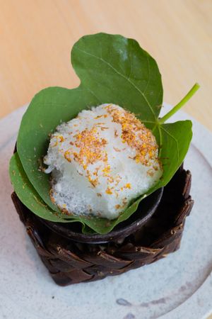 White rice ball garnished with spices and served on a green leaf in a woven basket vessel.