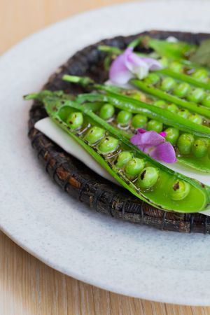 Vibrant green pea pods with purple flowers artfully arranged on dark textured plate.