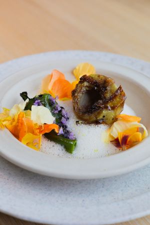 Elegantly plated dish with colorful garnishes, orange flowers and dark protein on a white ceramic plate.