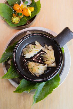 Steamed dumplings in dark broth served in a black ceramic pot with fresh green leaves and kumquats on the side.
