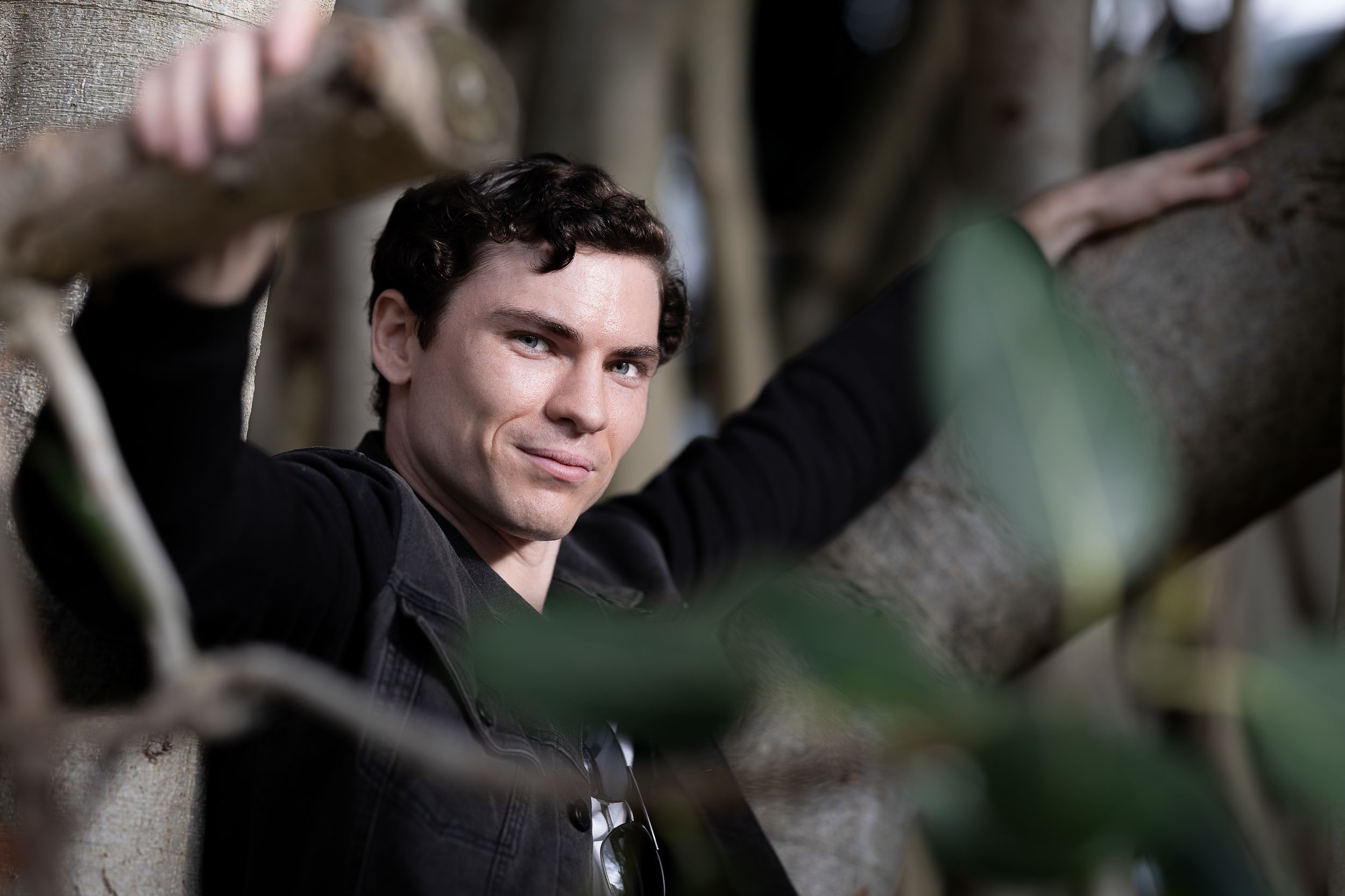 Young man with dark hair posing among tree branches, looking at camera in an outdoor setting.