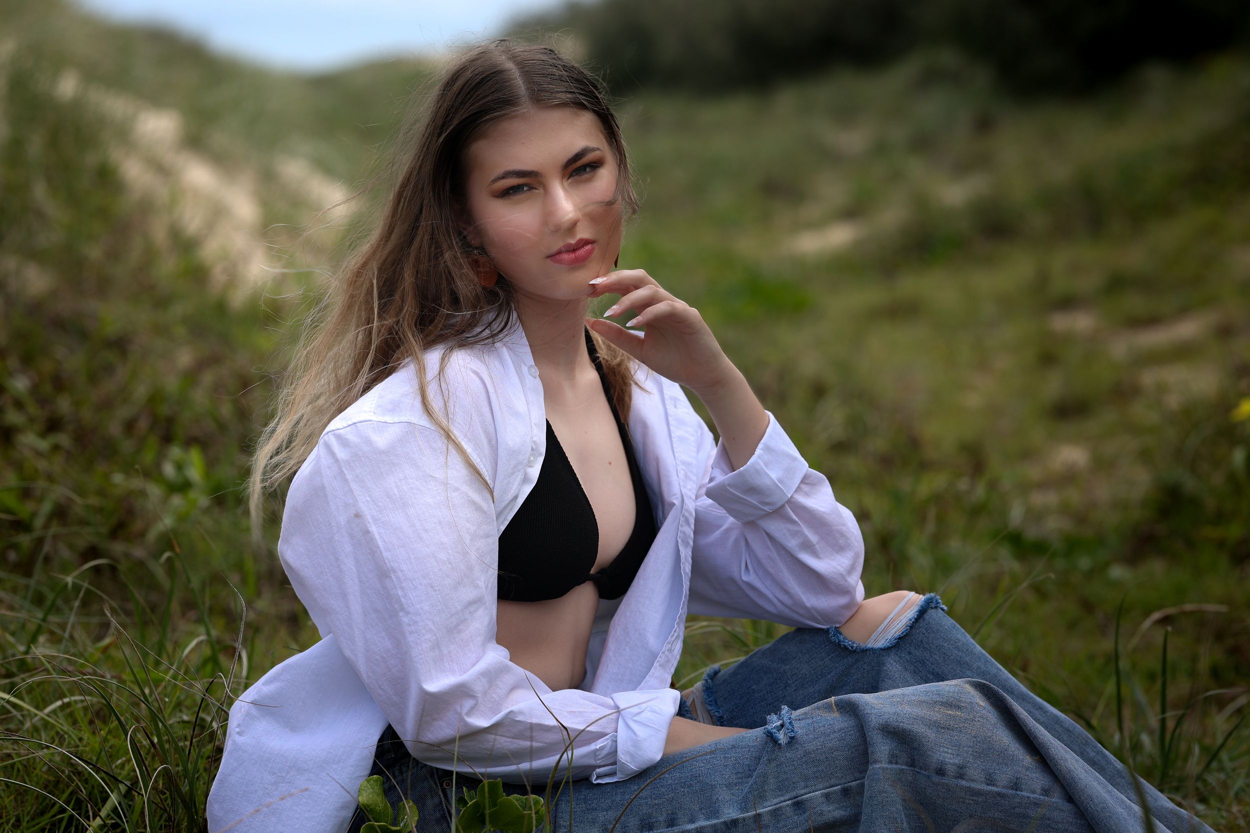 Young woman in white shirt and black bralette sits in grassy field, posing thoughtfully with hand near face.