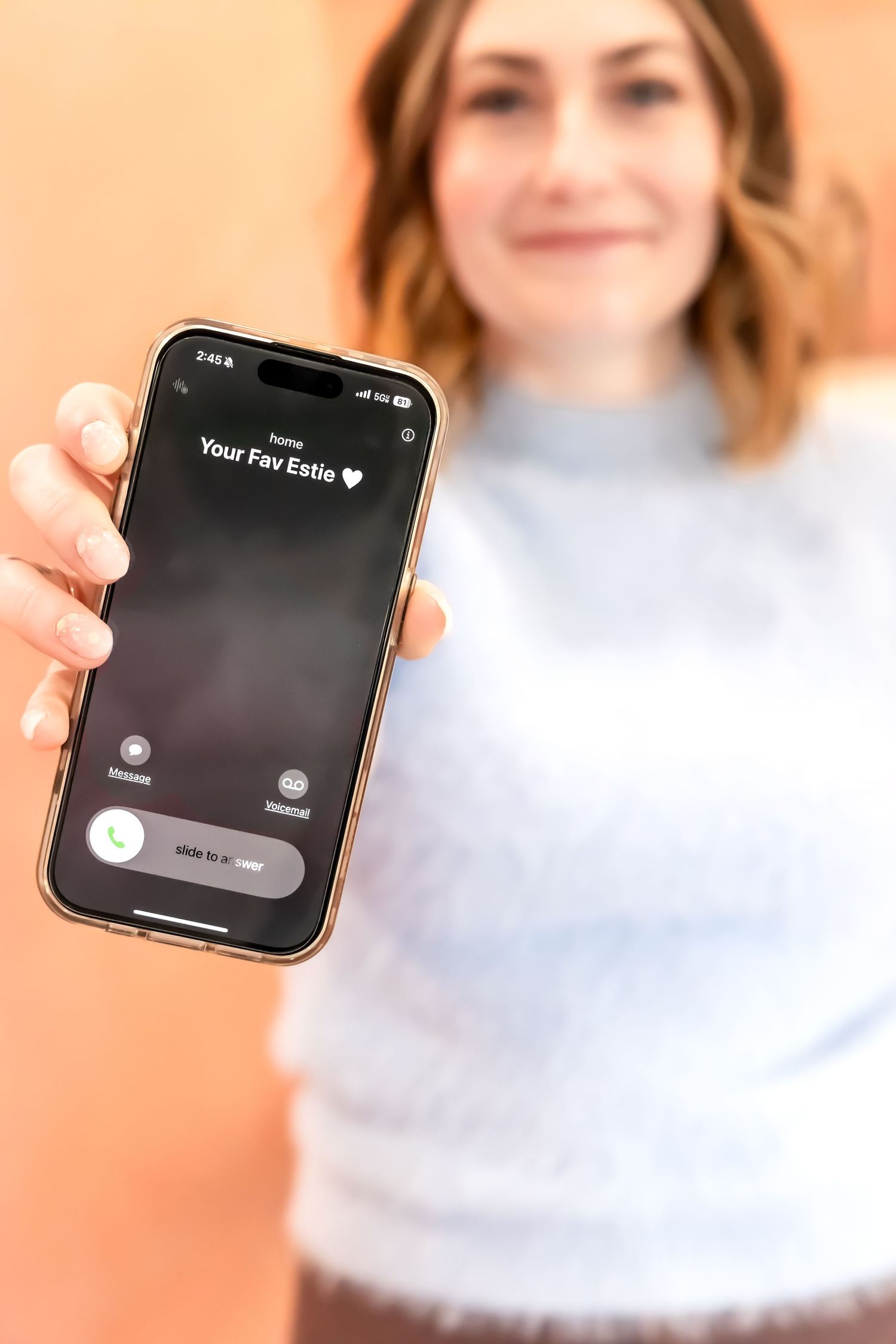 Someone wearing a white sweater holds up an iPhone displaying a dark app screen against a peach-colored background.