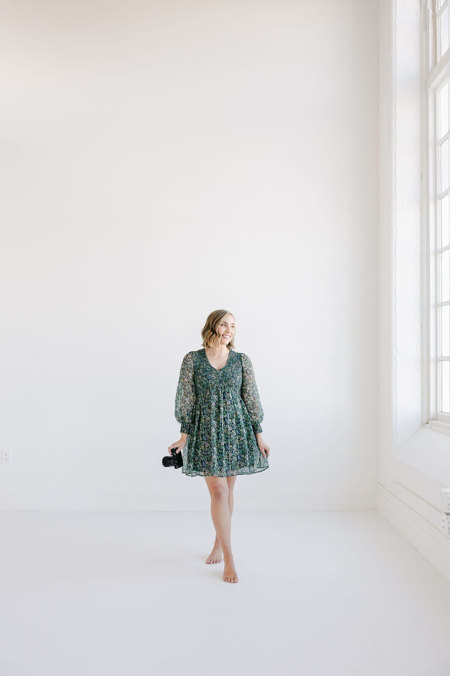 Woman in green floral dress posing barefoot against clean white studio backdrop.