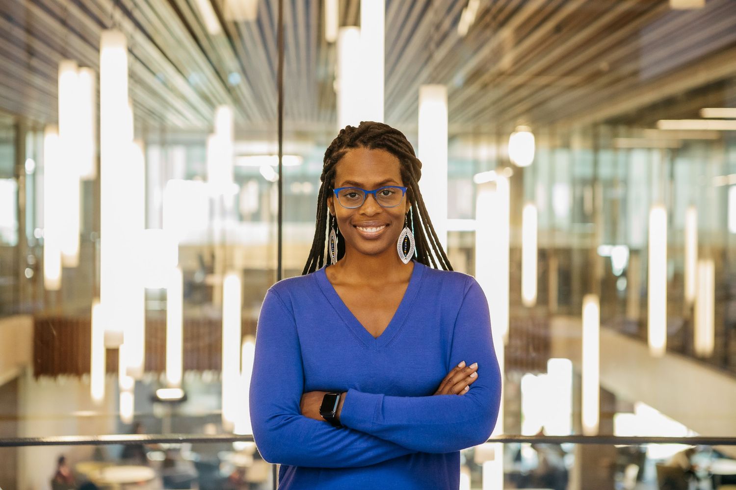 Professional in a bright blue sweater stands confidently with arms crossed in a modern office setting.