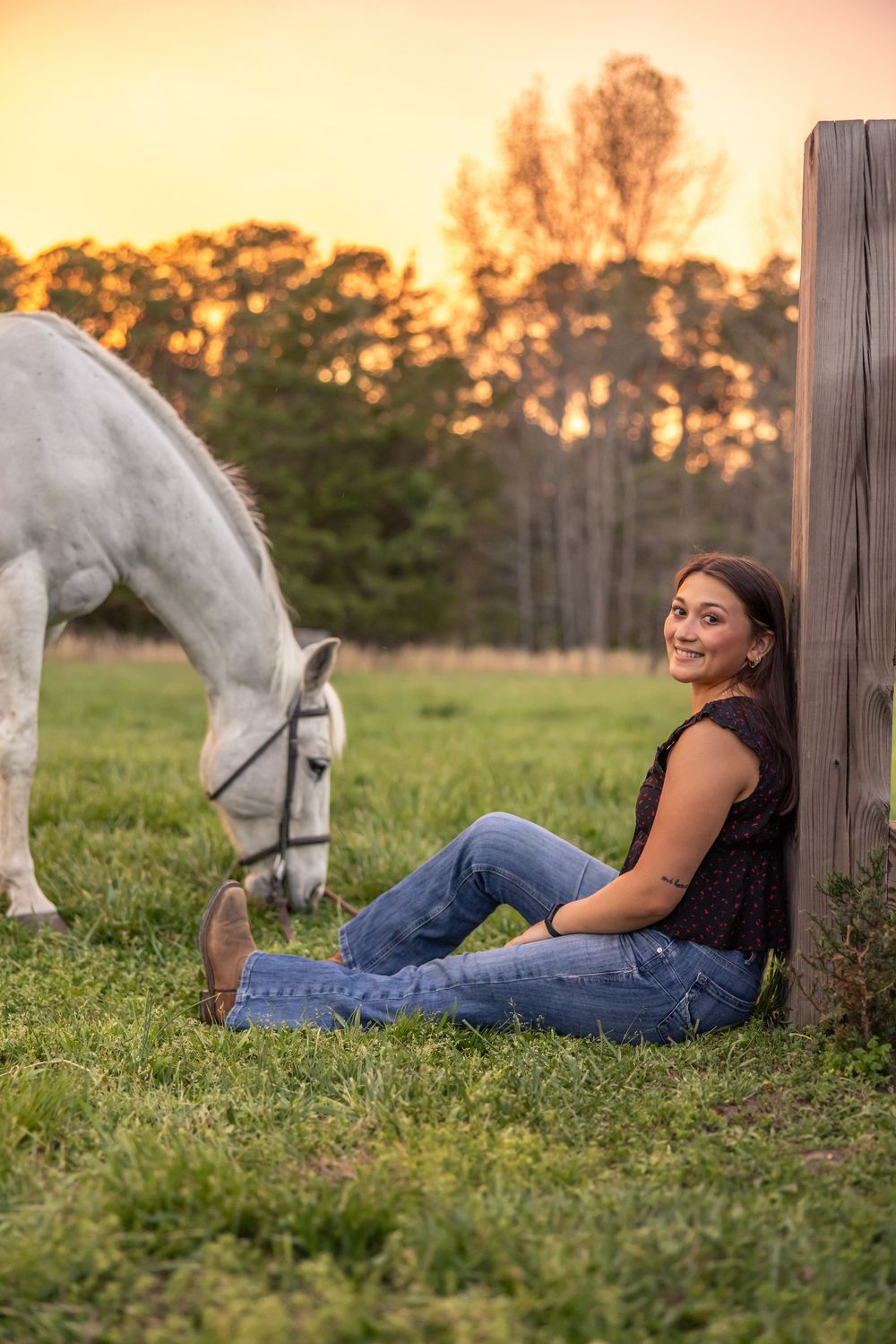 Young woman in jeans and boots sits smiling by a fence post beside a white grazing horse at golden sunset.