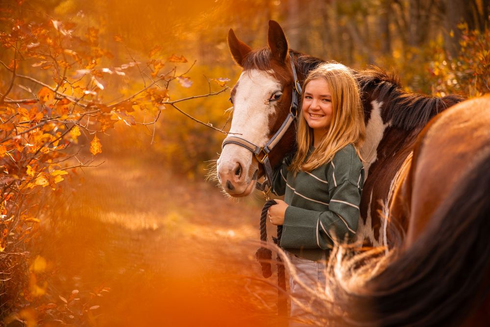 Smiling blonde girl embracing a brown and white horse surrounded by vibrant orange autumn foliage in a forest.