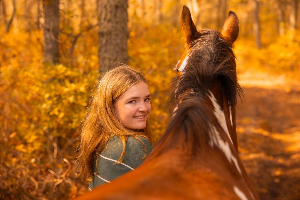 Smiling teenage girl with long blonde hair posing with a chestnut horse in an autumn forest setting.