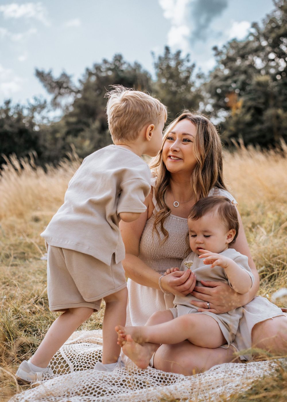 A heartwarming moment captured on a blanket in a sunny field with wheat grass and pine trees in the background. Family Photoshoot.
